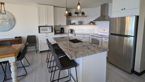 A modern kitchen with a central island, stainless steel appliances, and a dining area with a wooden table and bar stools. - Le Zéphyr - Waterfront (Beaulac-Garthby)