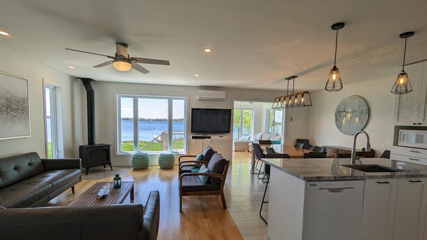 A modern living room with a large window, a fireplace, a sofa, and a dining area with a kitchen island. - Le Zéphyr - Waterfront (Beaulac-Garthby)
