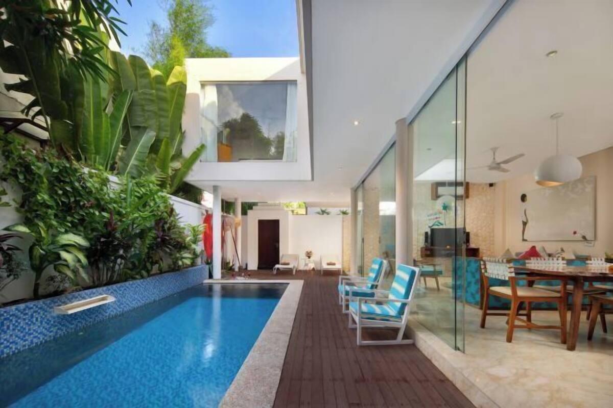 A modern pool area with a wooden deck, lounge chairs, and a glass wall leading to a dining area.