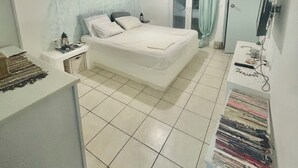 A bedroom with a bed, a desk, and a chair. There is a window with curtains, a ceiling light, and a wall-mounted TV. - Casa filareti (Ios)