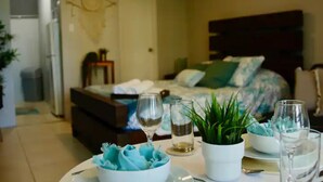 Un comedor ordenado con una mesa preparada para dos, una planta en maceta y una cama con cabecera al fondo.