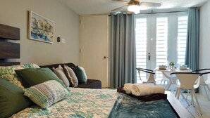Un dormitorio con cama, ventilador de techo, un cuadro, una mesa de comedor con sillas y una ventana con cortinas.