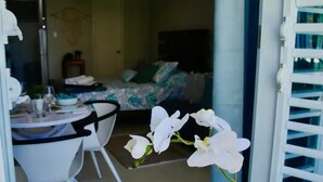 Una habitación con una cama, una mesita con sillas y un ventanal con persianas blancas.