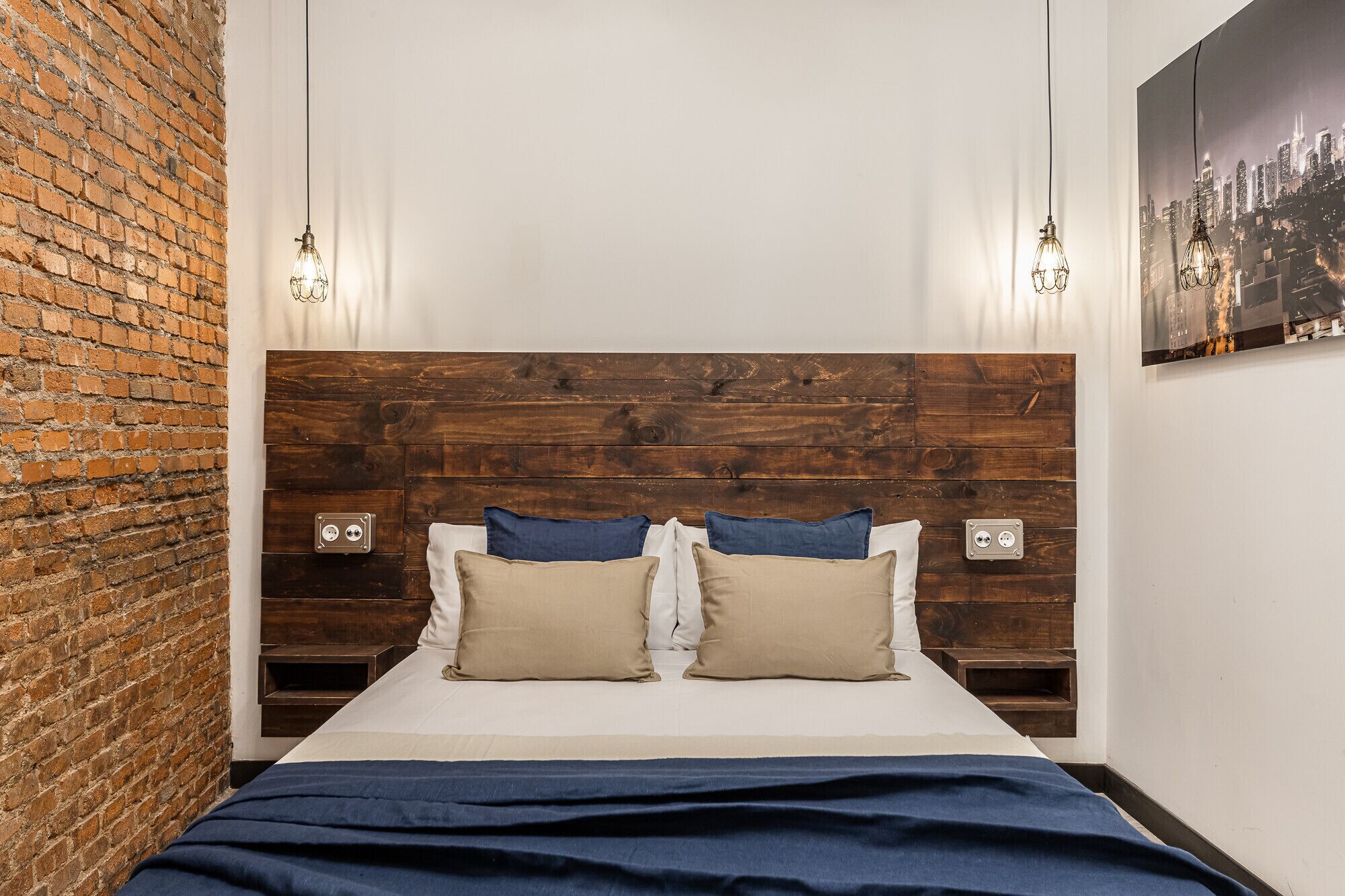 Una habitación de hotel con pared de ladrillo, cabecero de madera, dos mesitas de noche con lámparas, una cama con almohadas color beige y azul, y un cartel con vista a la ciudad en la pared.