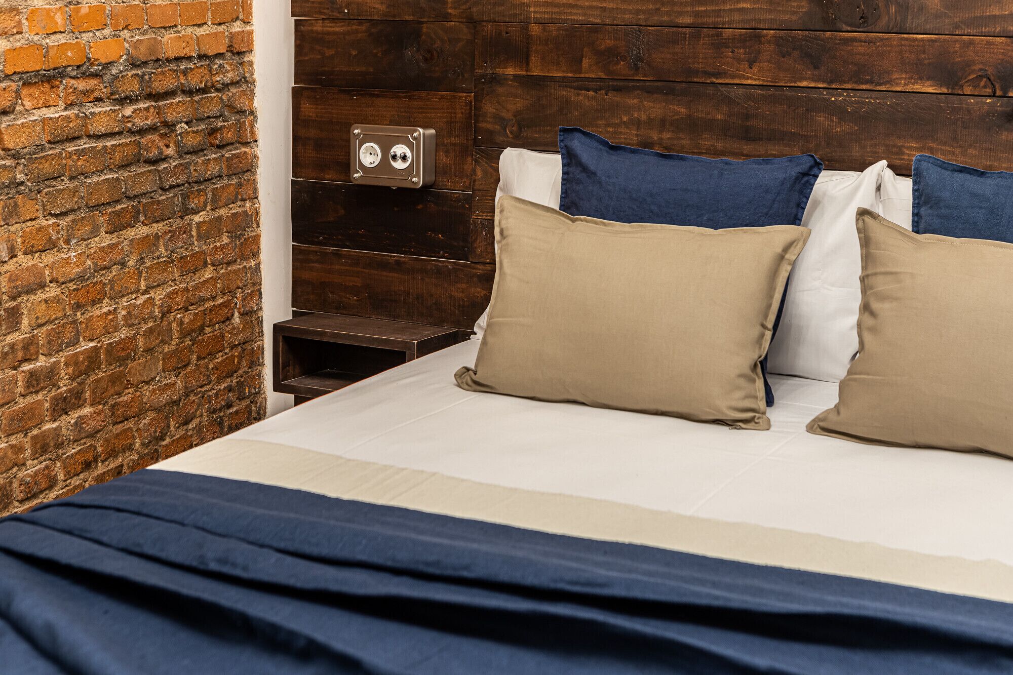 Een bed met een wit-blauwe spreidek en twee beige kussens, tegen een bakstenen muur.