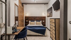 Een moderne slaapkamer met een accentmuur van baksteen, een houten hoofdbord, een bed met meerdere kussens, een bureau met koffiezetapparaat en een dressoir met lades genummerd 01-06.