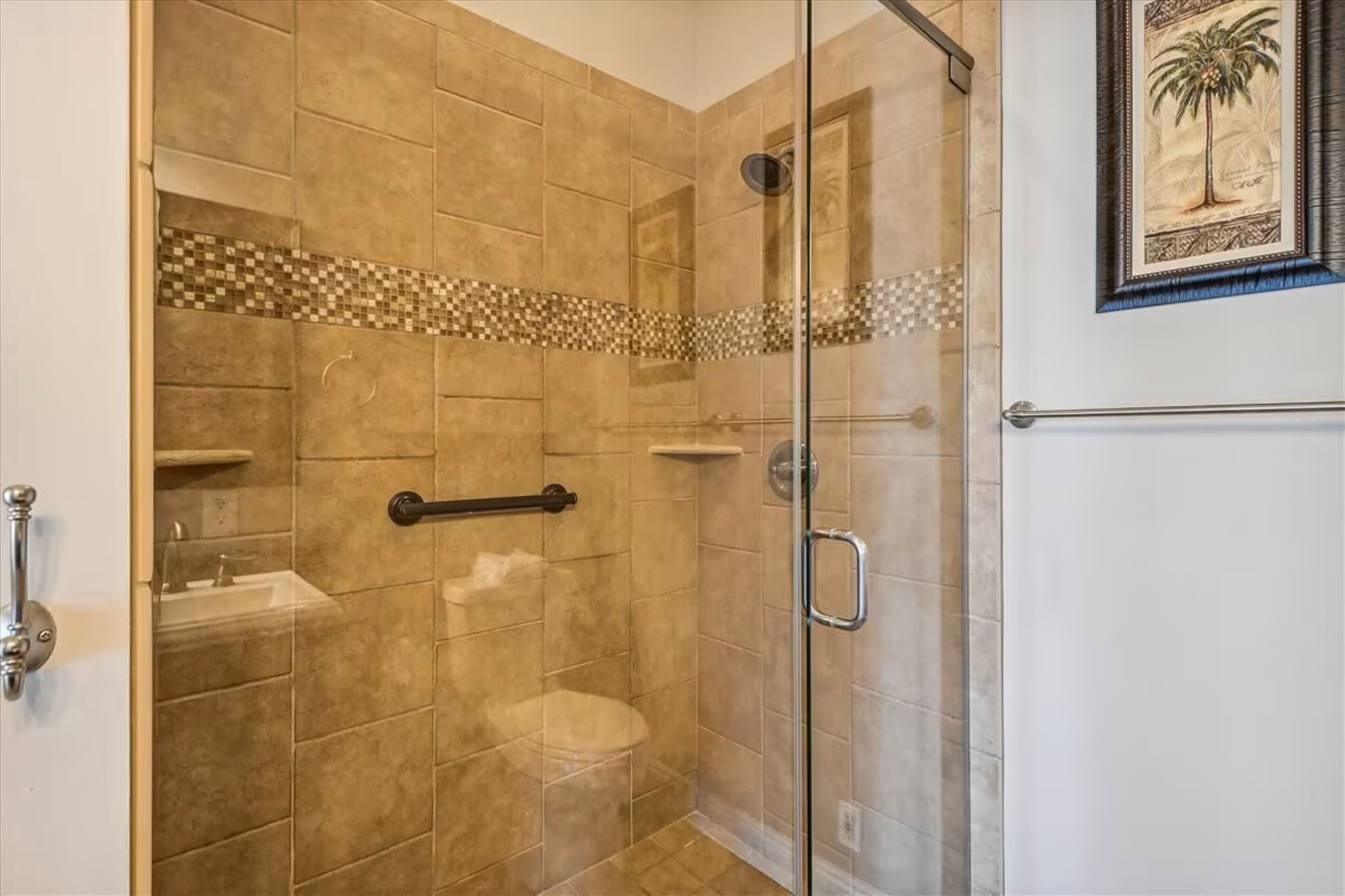 Une douche fermée par des parois en verre, avec des carreaux de couleur beige, une barre d’appui et un petit lavabo.
