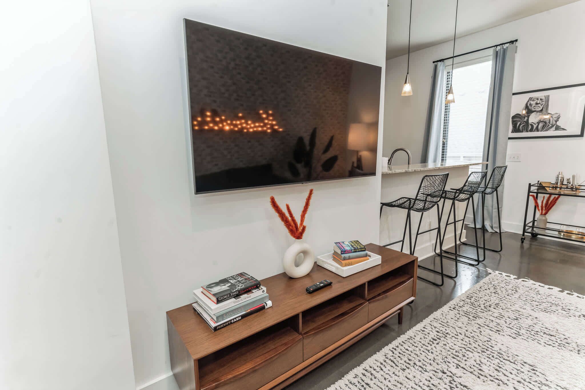 Una sala moderna con un televisor de pantalla plana grande montado en la pared, un mueble de madera con libros y un control remoto, y una zona de comedor con sillas y una mesa.