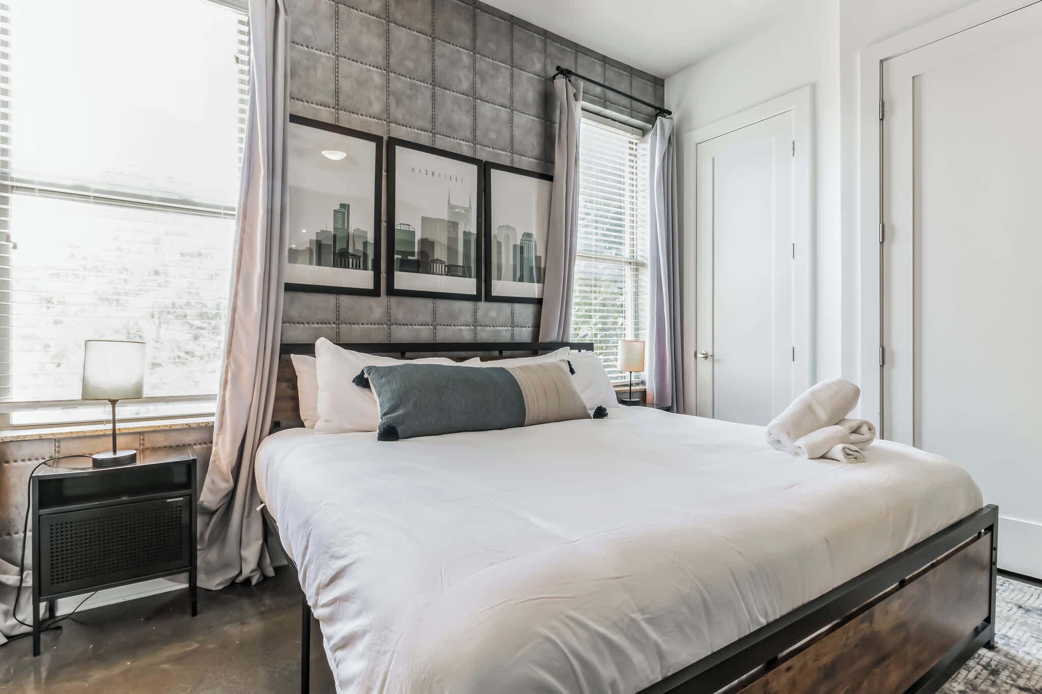 A neatly made bed with a headboard, a bedside table with a lamp, and a window with curtains.