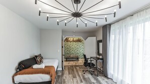 Une chambre moderne avec un lit, un bureau et une chaise, un luminaire au plafond original, et une vue sur une pièce dont le mur est décoré d’un motif.