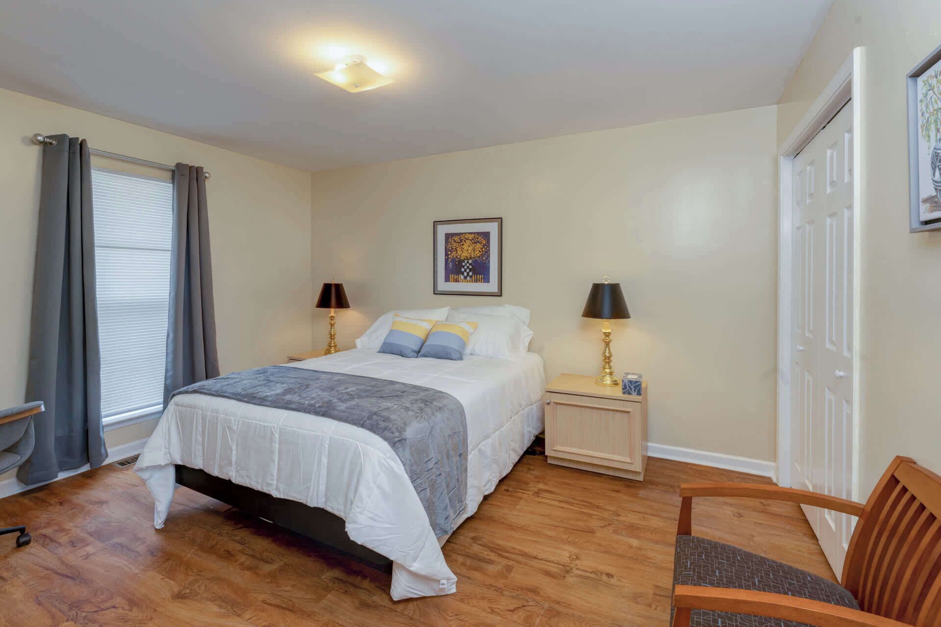 Un dormitorio con cama, dos mesitas de noche, una silla, ventana con cortinas, un cuadro en la pared y una puerta.