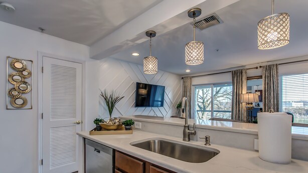 A modern kitchen with a stainless steel sink, wooden cabinets, pendant lights, and a large window with curtains. - Riverfront Condo Minutes from Broadway (Nashville)
