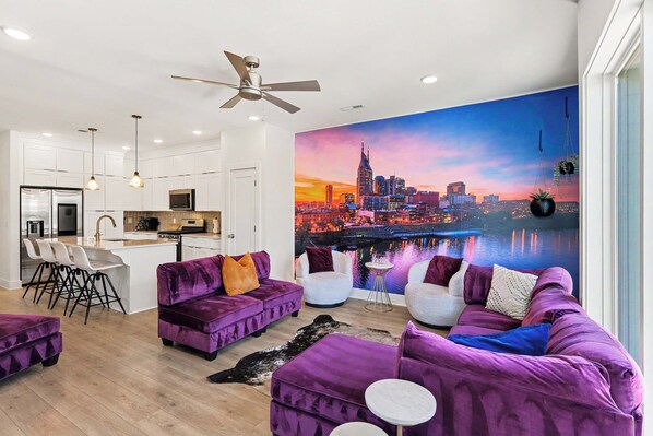 Una sala moderna con sofás morados, un mural con un paisaje urbano y una zona de cocina.