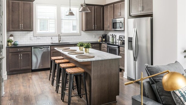 Una cocina moderna con armarios de madera oscura, una isla central con taburetes, electrodomésticos de acero inoxidable y una lámpara colgante.