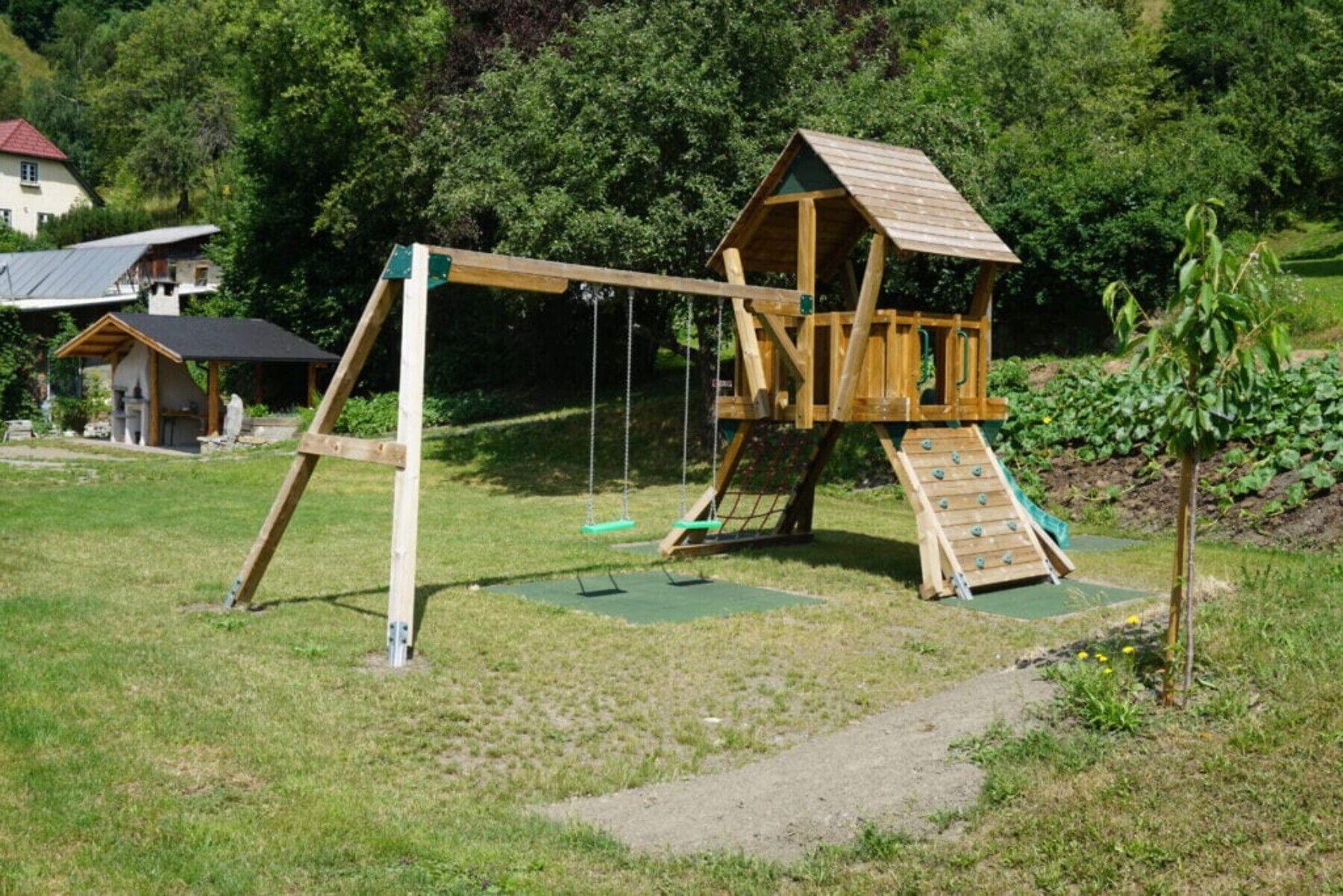 Un conjunto de columpios de madera con pared de escalar y tobogán, ubicado en un área verde con una casa al fondo.