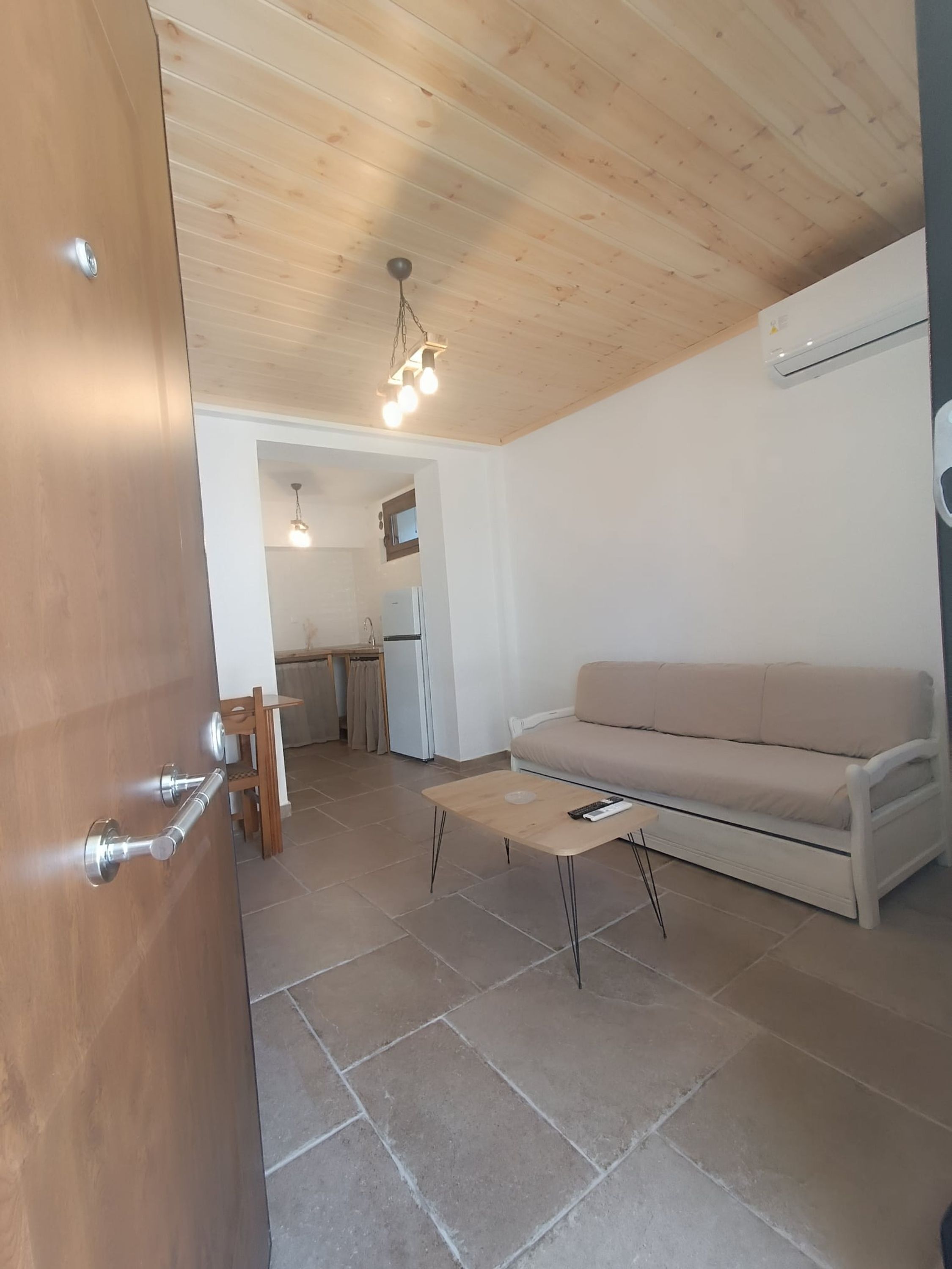 Een moderne woonkamer met een houten plafond, een beige bank, een kleine salontafel, en een keuken in de achtergrond.