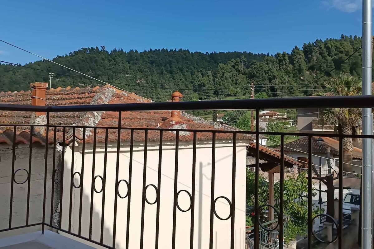 Un balcón con barandilla de metal que da a una zona residencial con tejados de teja y una colina boscosa al fondo.