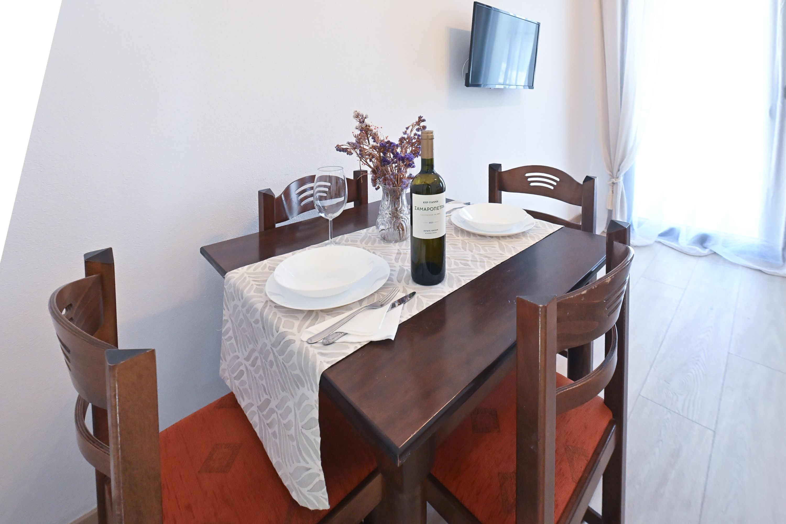 Un tavolo da pranzo con una bottiglia di vino, un vaso con fiori e una TV a schermo piatto fissata al muro.