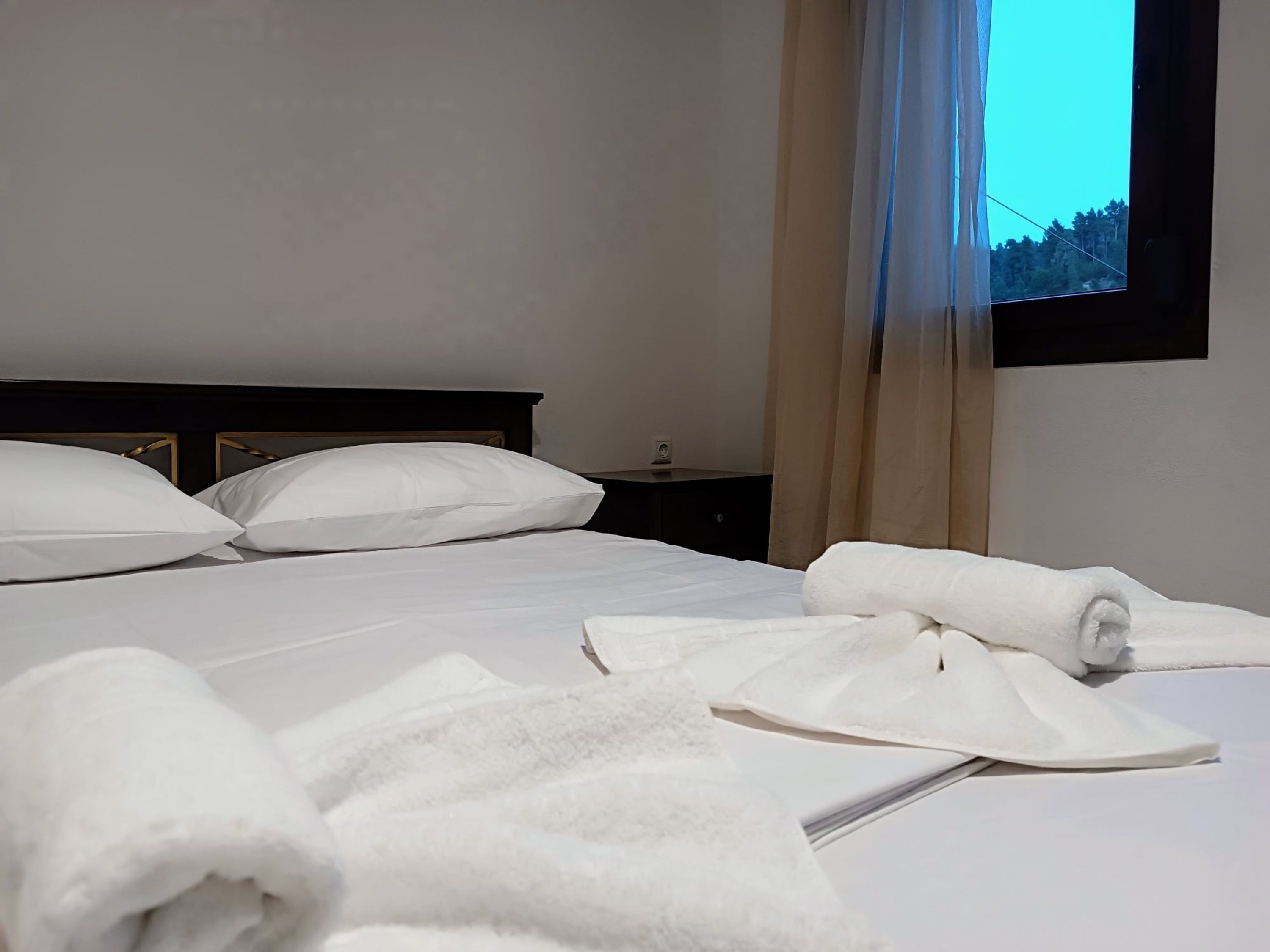 A neatly made bed with white linens and pillows, a bedside table with a lamp, and a window with curtains.