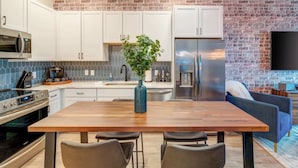 Una cocina moderna con armarios blancos, una pared de acento de ladrillo, un refrigerador de acero inoxidable y un comedor con mesa y sillas de madera.