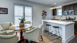 A modern kitchen with a central island, bar stools, and a dining area with a round table and chairs. - Quiet Condo l Prime Locale & mins to DT (Nashville)