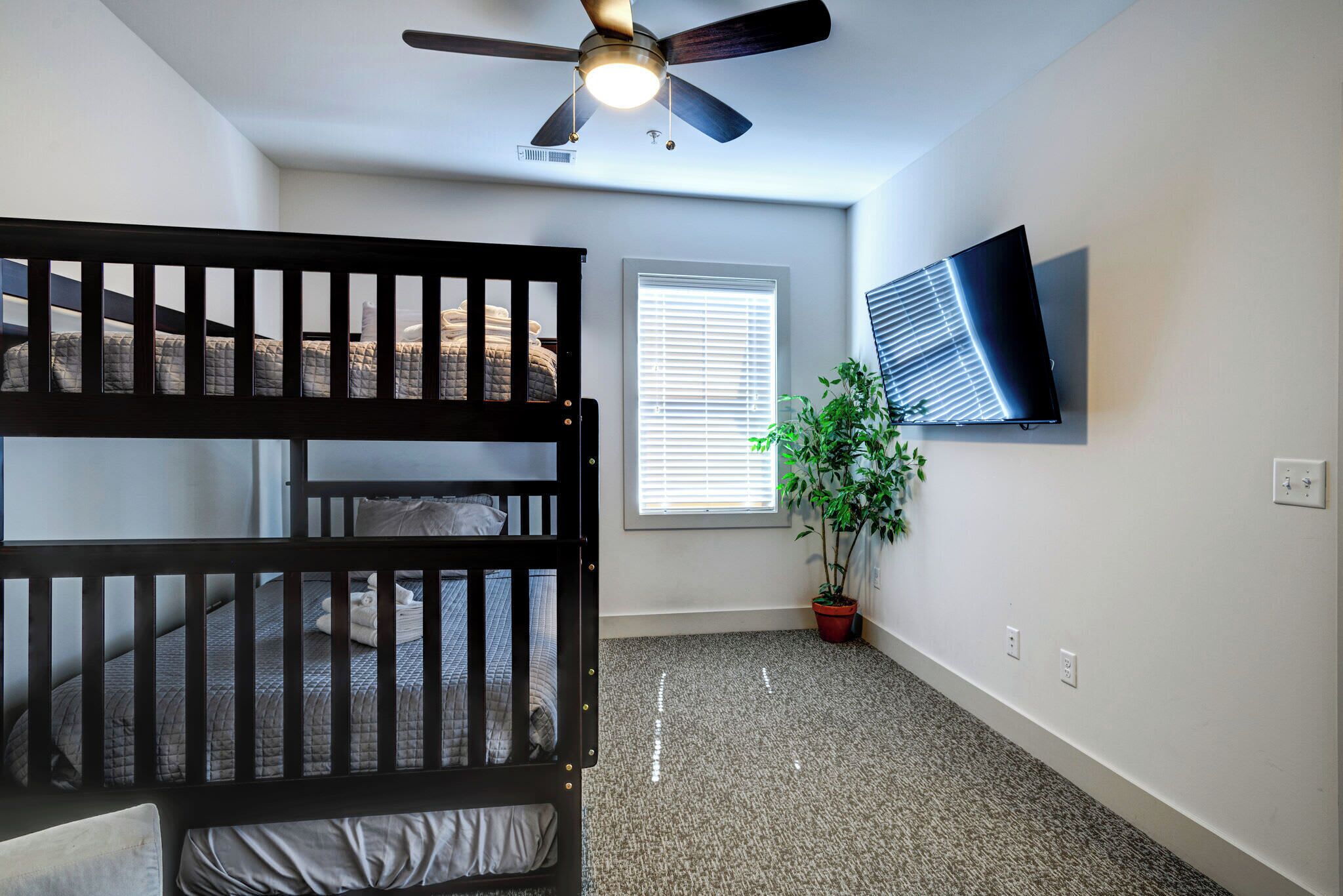 A bedroom with two bunk beds, a ceiling fan, a TV, a window with blinds, and a potted plant.
