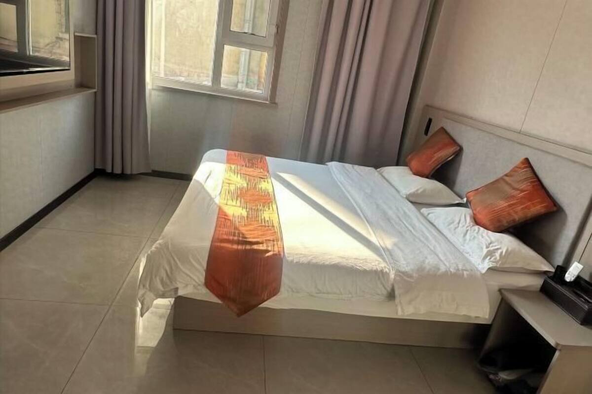 Een net opgemaakt bed met een witte dekbedovertrek en een oranje plaid, een nachtkastje met een lamp, en een raam met gordijnen.