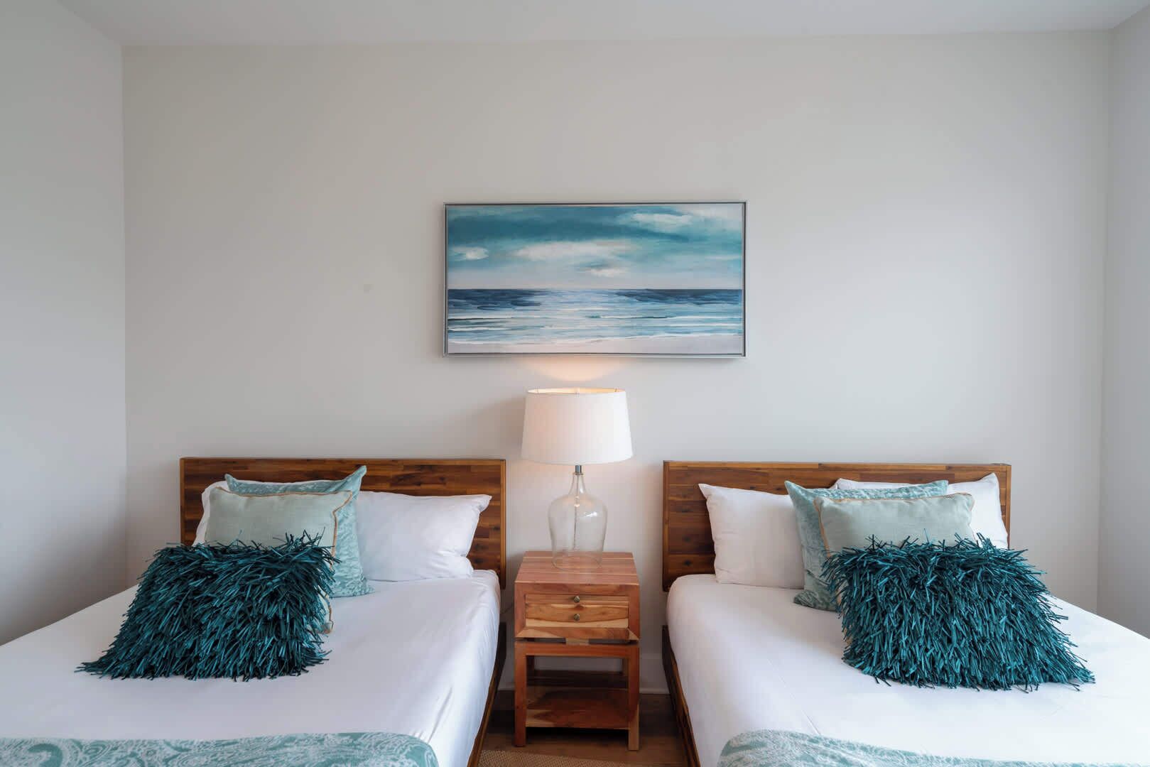 A room with two beds, each with a wooden headboard, a nightstand with a lamp, and a painting of a seascape on the wall.