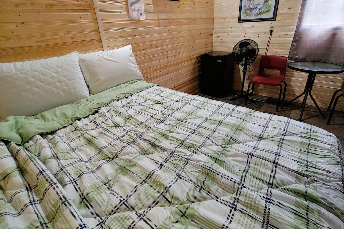 Une chambre avec un lit, une petite table, une chaise, un ventilateur et un panneau « Il est interdit de fumer » sur le mur.