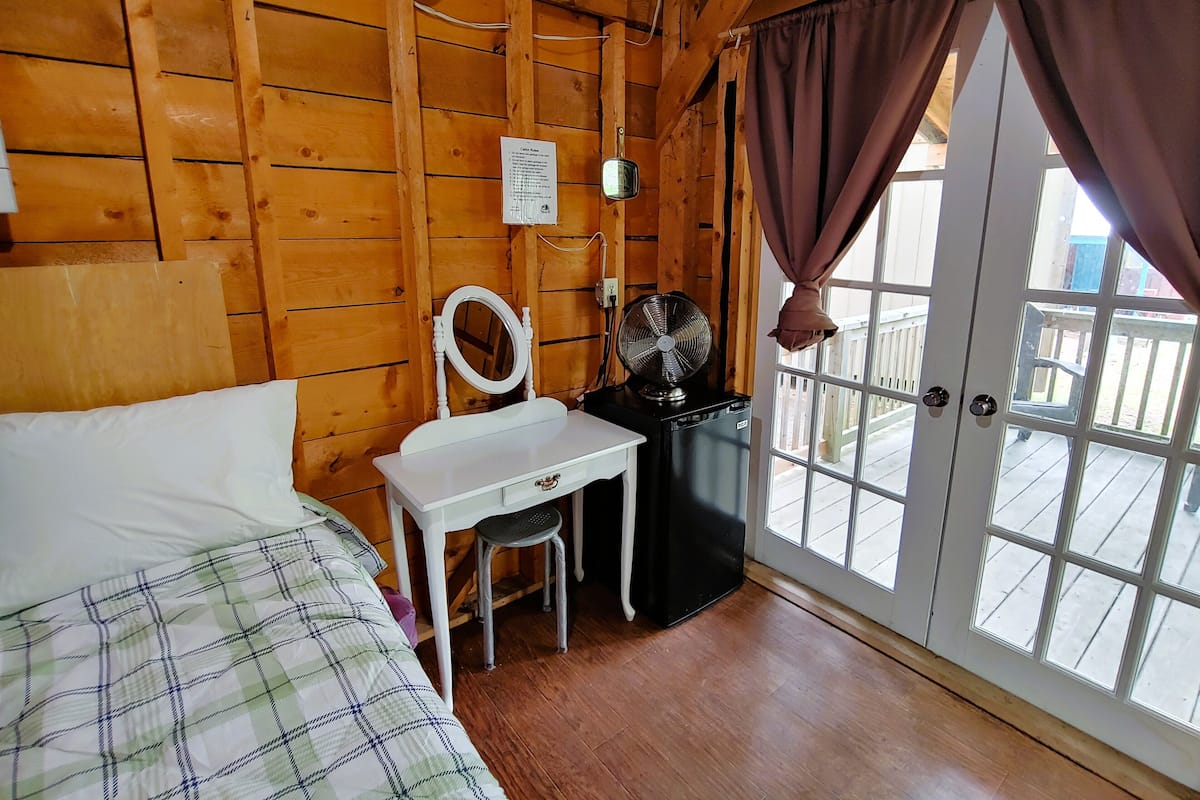Une chambre dans un chalet en bois, avec un lit, un petit bureau doté d’un miroir, un ventilateur et une fenêtre avec des rideaux.