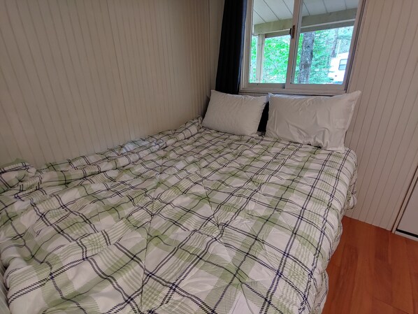 A bed with a checkered bedspread, a window with a view of greenery, and a wooden floor.