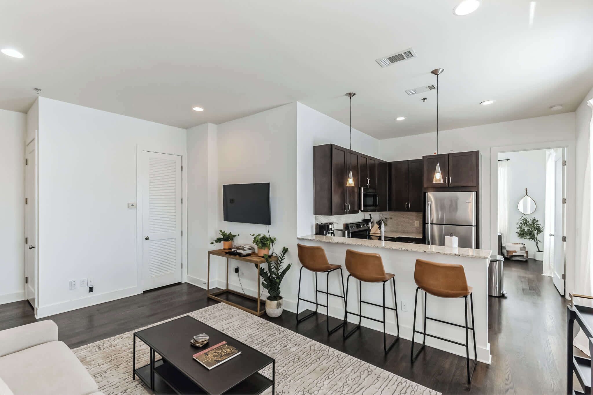 Un appartement moderne comprenant un salon, une cuisine et un coin repas. Il est équipé d’un téléviseur à écran plat, d’un îlot de cuisine avec des tabourets et d’une table à manger.
