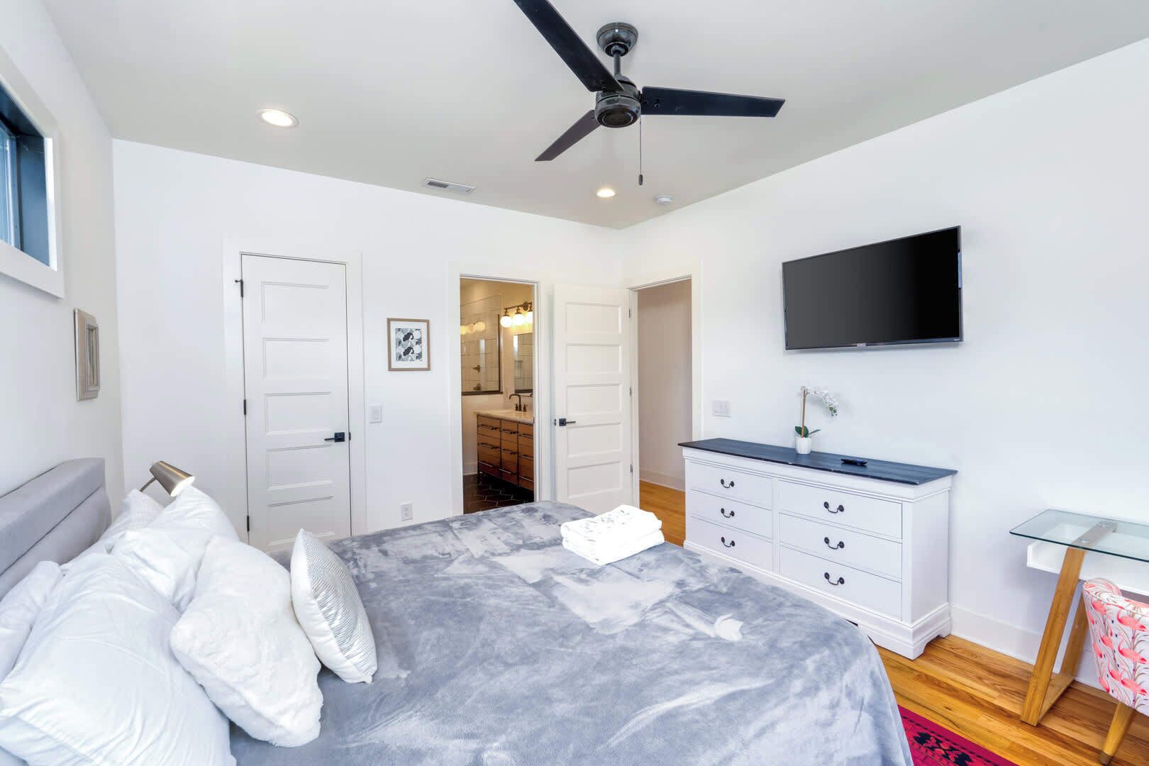 A bedroom with a bed, a ceiling fan, a TV, a dresser, and a bathroom visible through an open door.