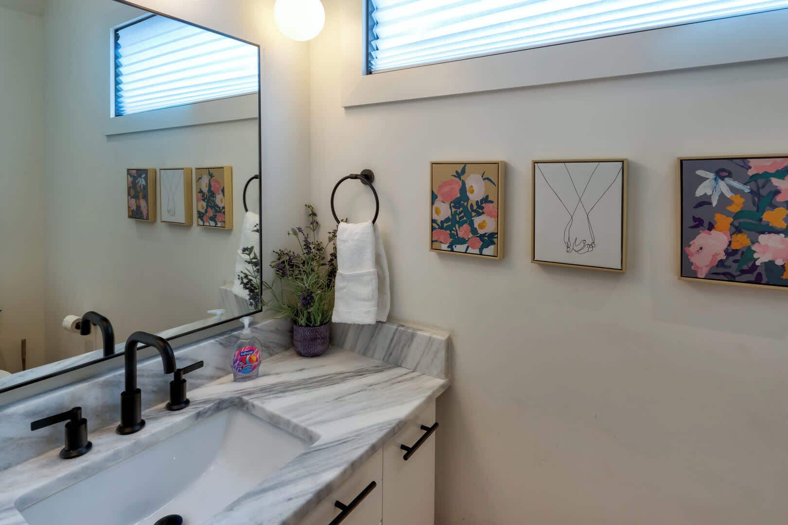A bathroom with a marble sink, black faucets, a mirror, and wall-mounted towel rings. There are framed floral artworks on the wall.