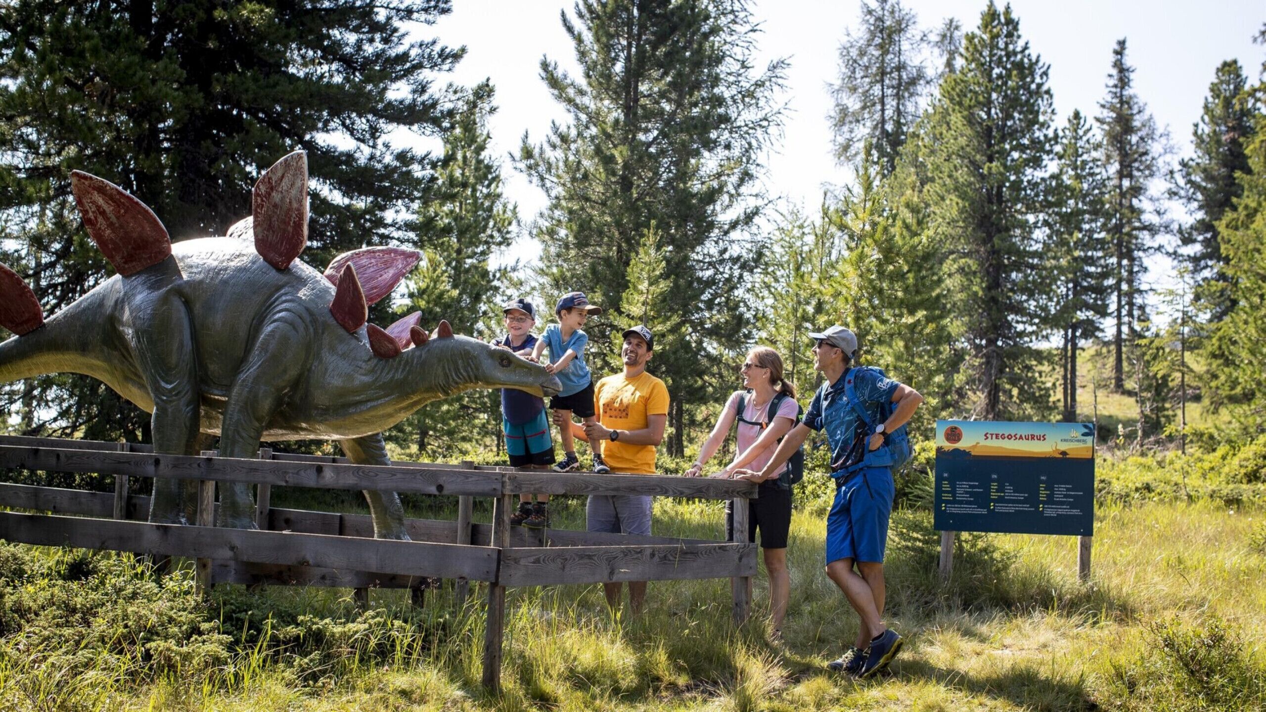A group of people observing a dinosaur exhibit in a natural setting with a wooden fence and informational sign.