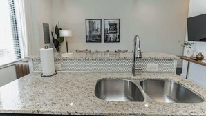 A modern kitchen with a granite countertop, stainless steel sink, and a backsplash with a geometric pattern. There are two framed pictures on the wall, a potted plant, and a vase on the counter. - Couple's Retreat Near Vanderbilt University! (Nashville)