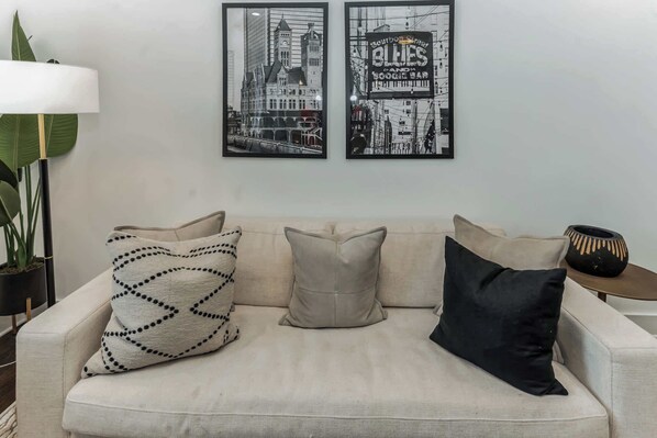 Una sala de estar con un sofá, tres almohadas, una lámpara de pie, una planta en maceta y dos cuadros en la pared.