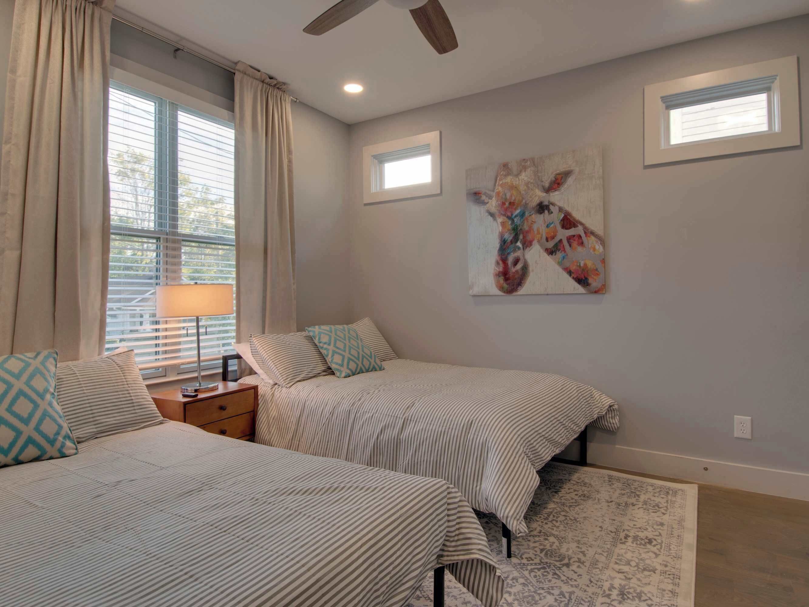Une chambre avec deux lits, un ventilateur de plafond, une fenêtre avec des stores, une lampe et un tableau représentant une girafe accroché au mur.