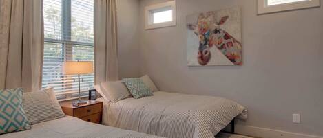 Une chambre avec deux lits, un ventilateur de plafond, une fenêtre avec des stores, une lampe et un tableau représentant une girafe accroché au mur.