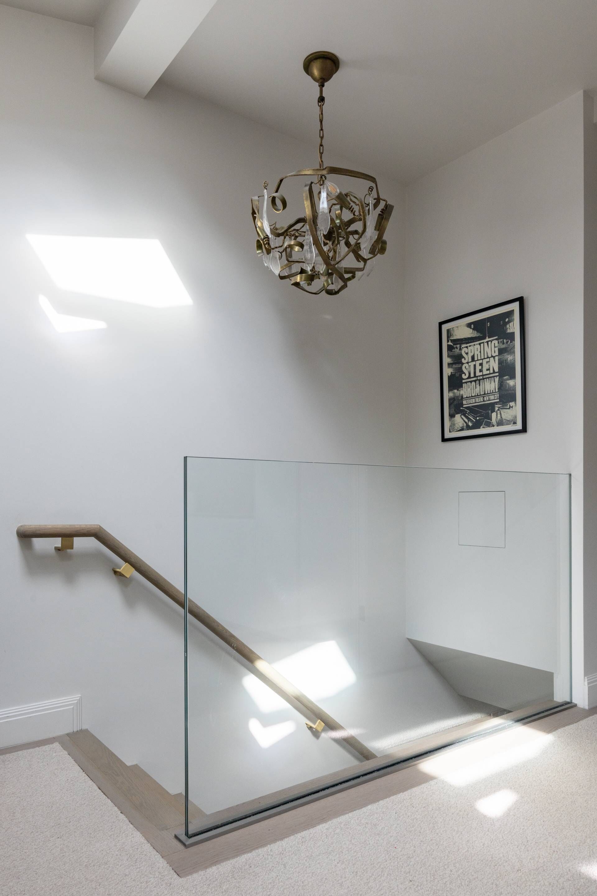 Un escalier moderne avec des rampes en verre et une main courante en bois, un lustre et une affiche encadrée au mur.