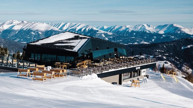 Un moderno edificio de estación de esquí con fachada de vidrio, rodeado de montañas cubiertas de nieve.