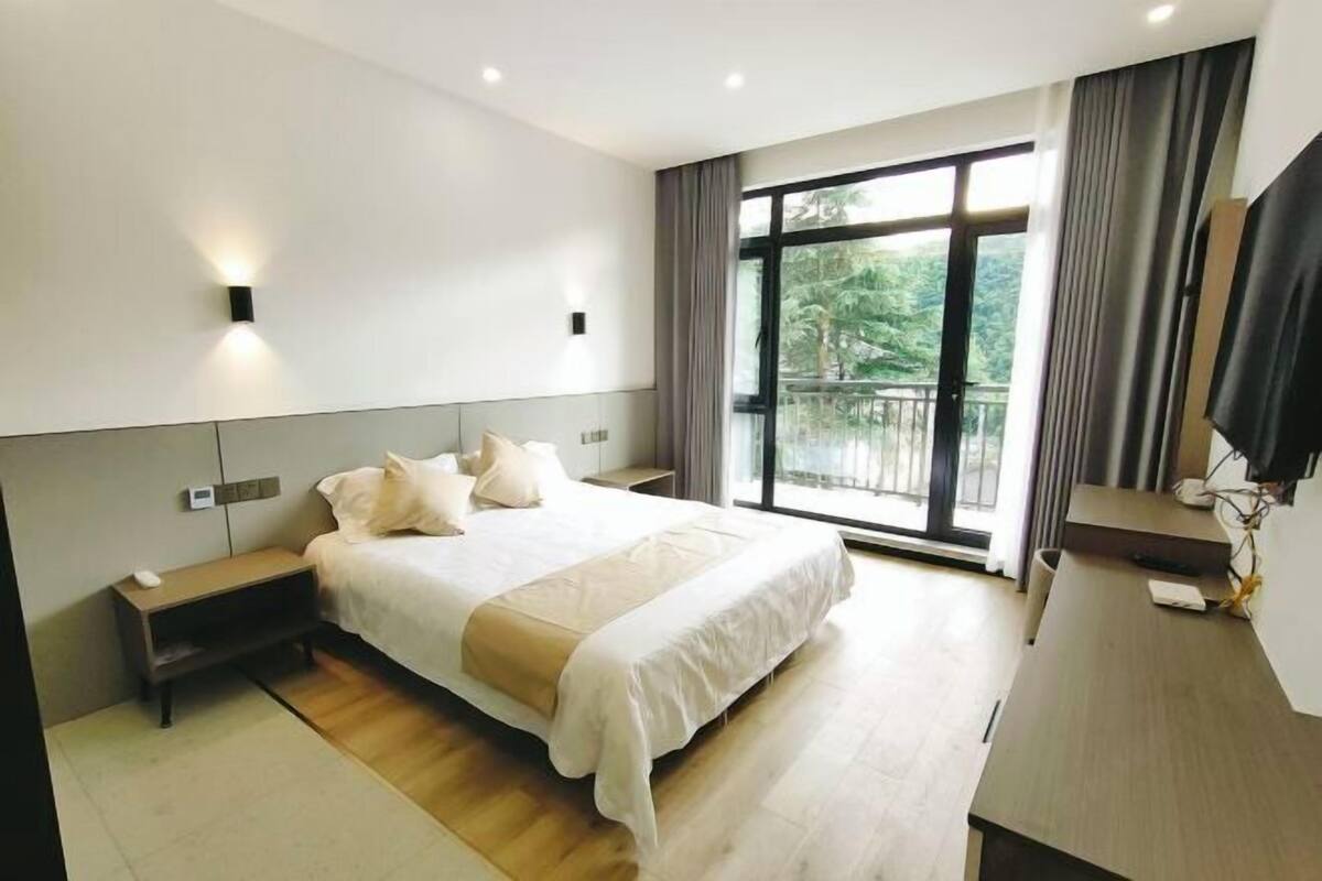 A modern hotel room with a large bed, bedside tables, a desk, and a balcony with a view of greenery.