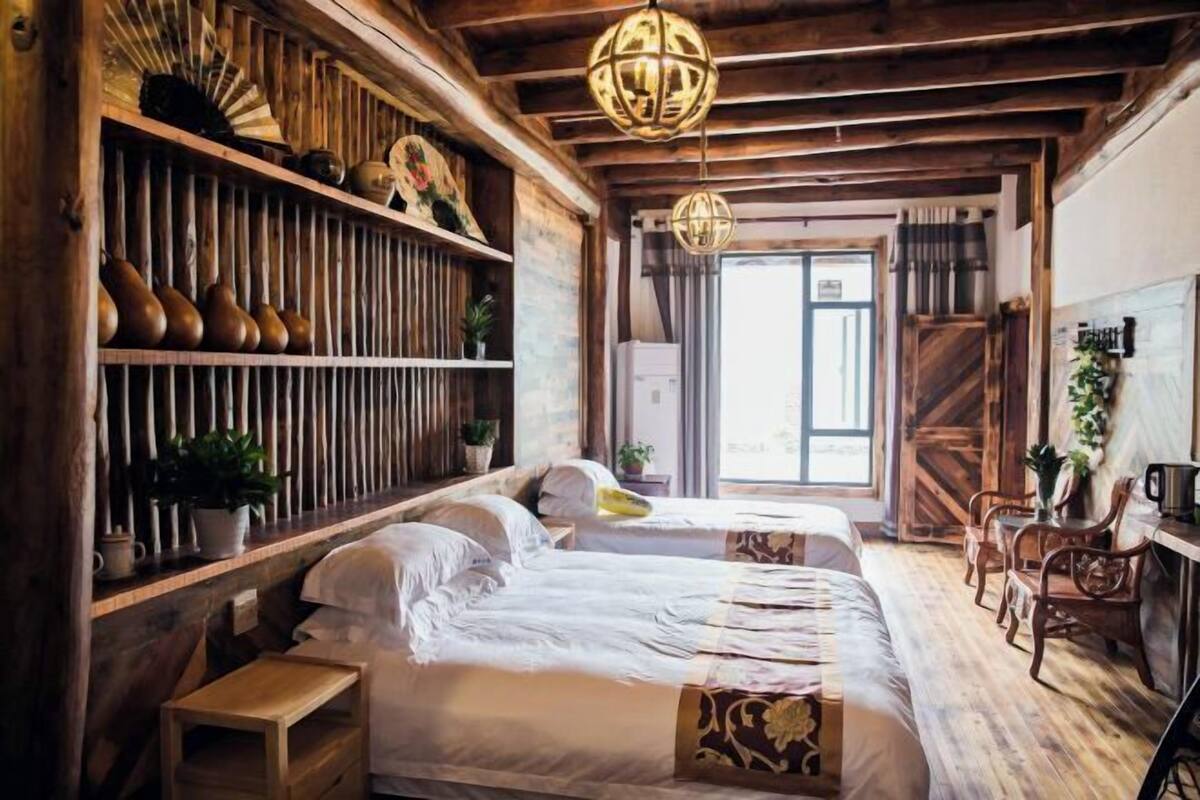 A rustic room with wooden beams, a large shelf with decorative items, two beds, a desk, and a window with curtains.