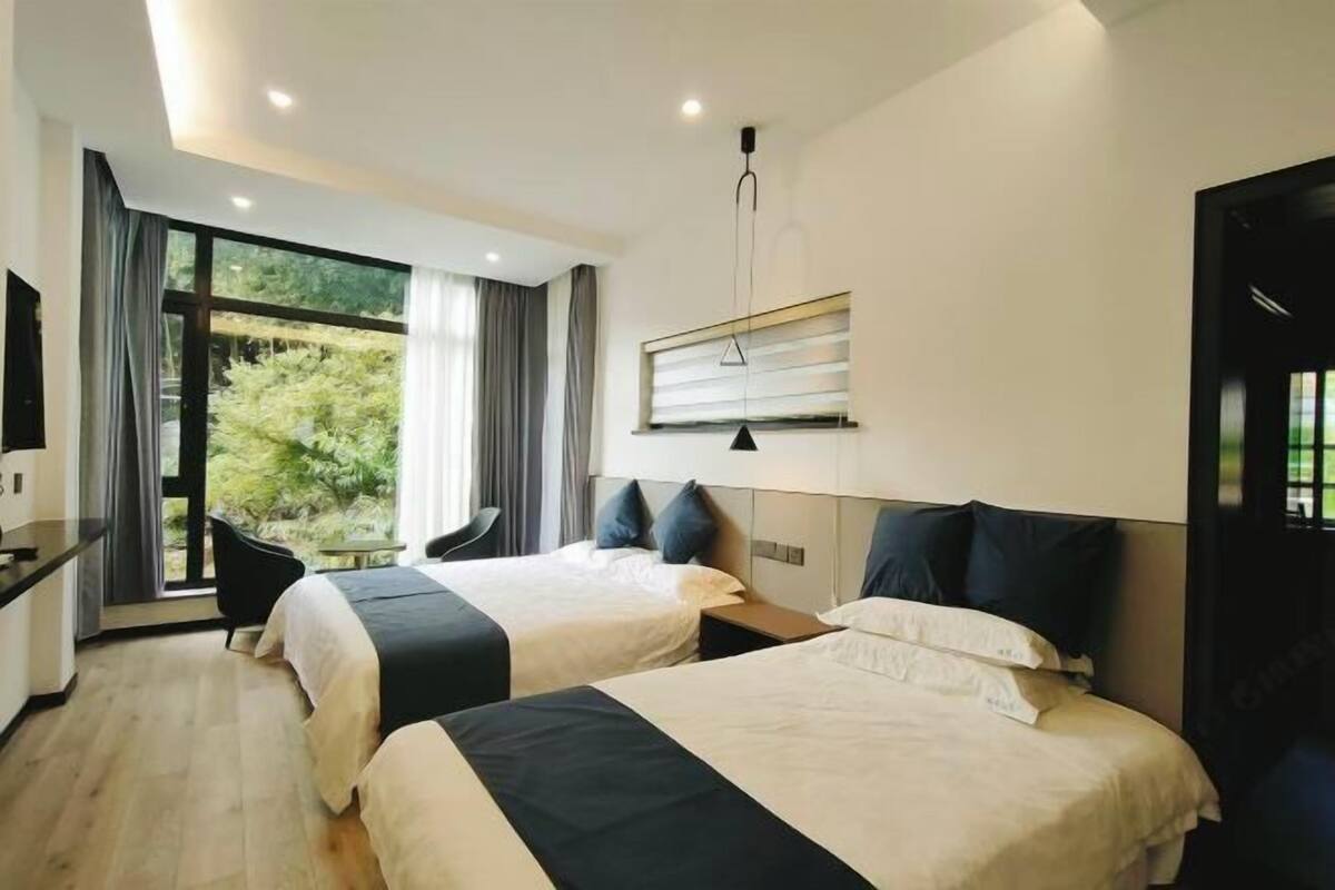 A modern hotel room with two beds, a desk, and a large window offering a view of greenery.