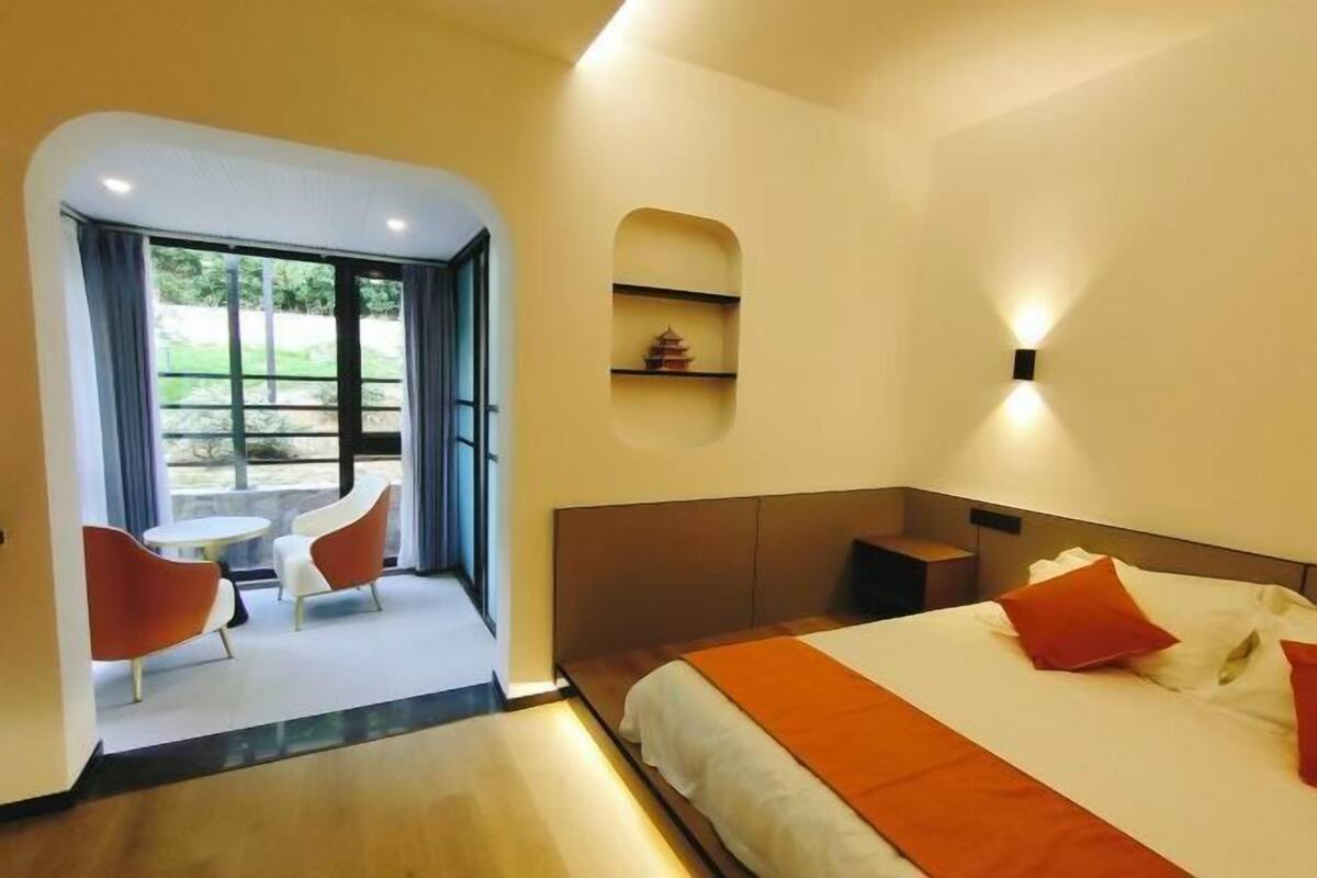 A modern hotel room with a bed, a small table, two chairs, a window, and a wall-mounted shelf.