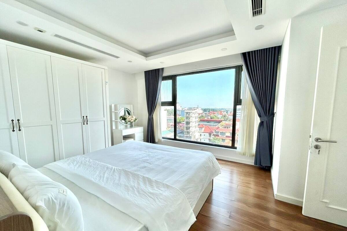 A modern bedroom with a large bed, white wardrobe, and a city view through a large window.