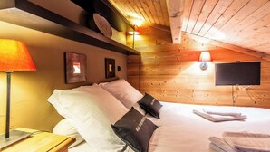 A cozy bedroom with a wooden ceiling, a bed with white and black pillows, a bedside lamp, a flat-screen TV, and a shelf with decorative items. - Slow Chalet MtBlanc Soleil Ski-in/out Nice View Balcony Elevator Parking (Manigod)