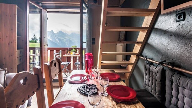 A dining area with a table set for two, a view of mountains, and a wooden interior. - Slow Chalet MtBlanc Soleil Ski-in/out Nice View Balcony Elevator Parking (Manigod)