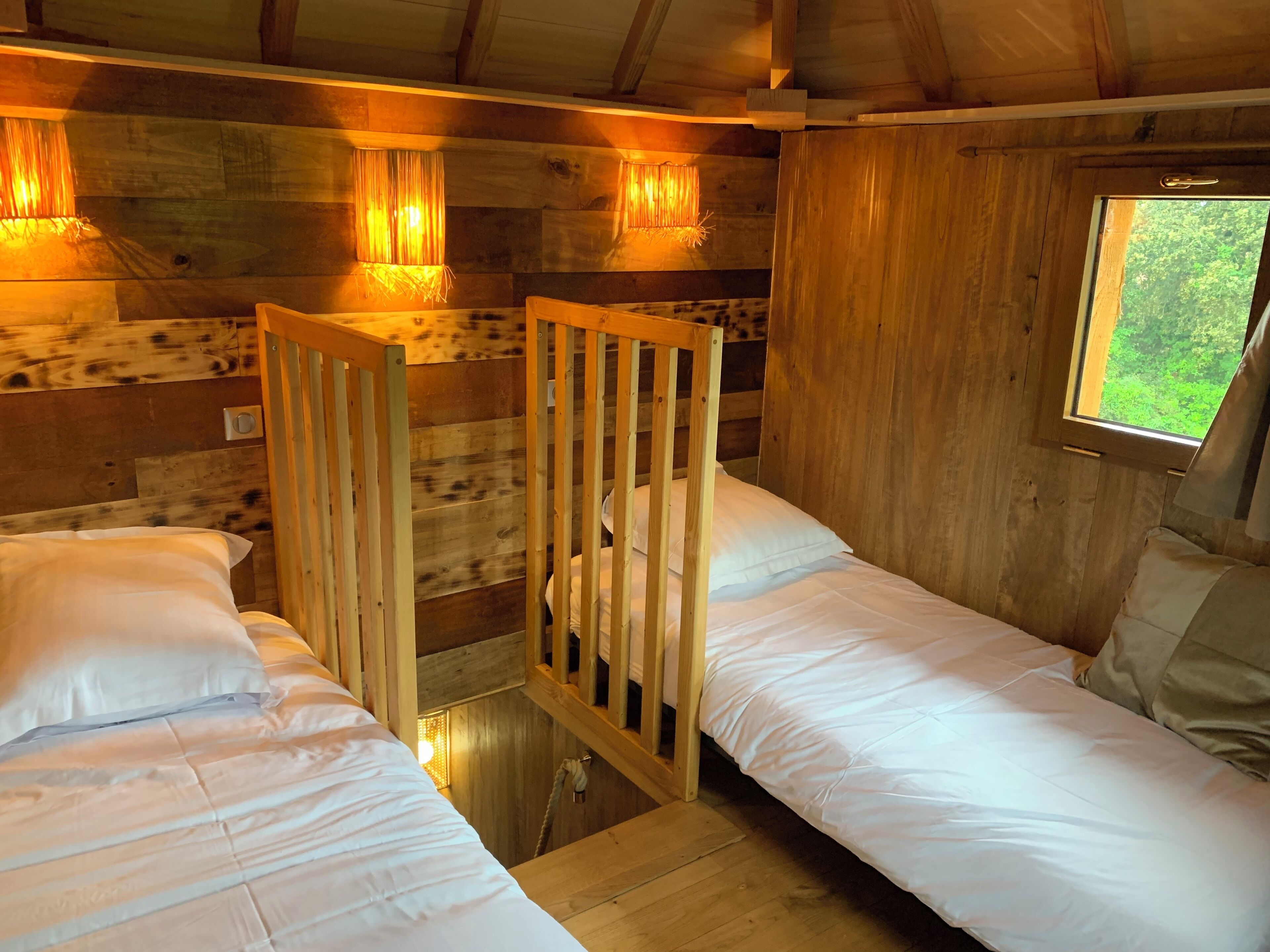 Eine Hütte mit zwei Holzbettstellen, einem Fenster mit Blick auf Grünflächen und Wandleuchten.