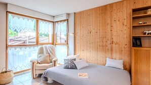 Une chambre à coucher avec un lit, une chaise, une armoire en bois et une vue sur un immeuble à travers de grandes fenêtres.